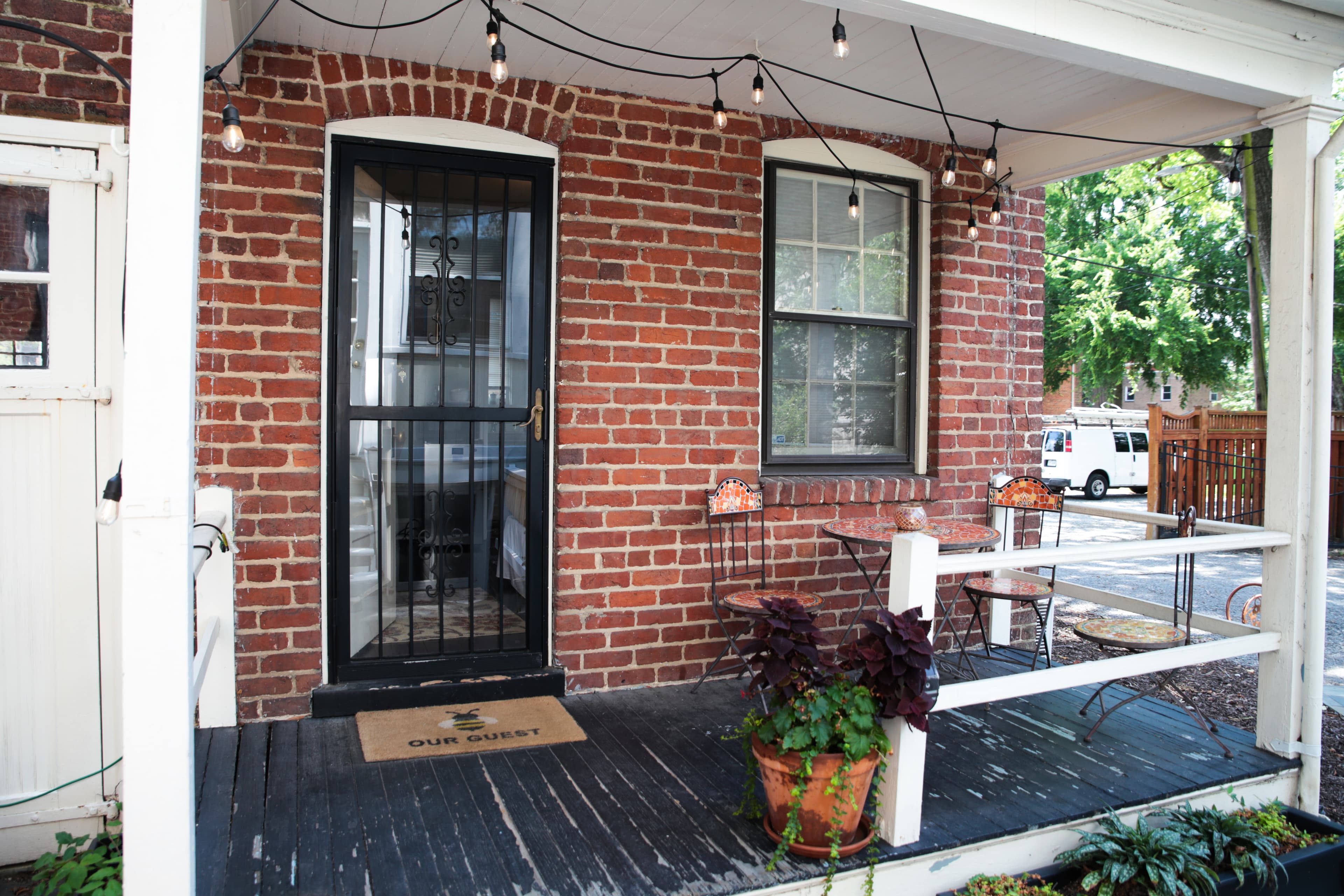 The exterior of a brick carriage house with a covered porch and a patio with outdoor furniture and potted plants, surrounded by lush greenery.