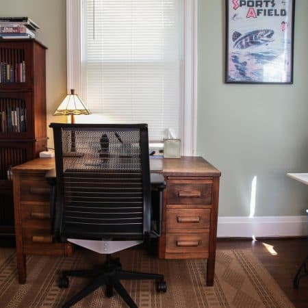 An office with light green walls, a wooden desk with a black mesh office chair, and a window with blinds.