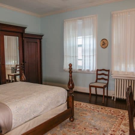 A bedroom with light blue walls, a dark wood four-poster bed, a large wooden armoire, and two windows with white curtains.