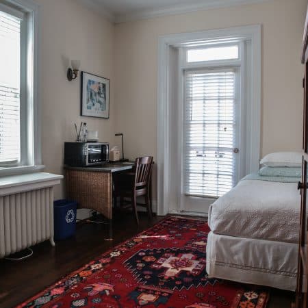 A bedroom with a bed, a desk with a microwave, a red patterned rug, and a window with blinds.