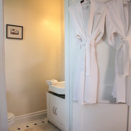 A bright bathroom with white subway tiles, a pedestal sink, a toilet, and a window with white curtains.