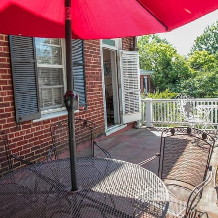 A brick patio with a black metal table and chairs, shaded by a red umbrella, and a white railing.