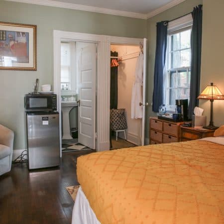 A bedroom with light green walls, a bed with a yellow comforter, a mini-fridge and microwave, and open doors to a bathroom and closet.
