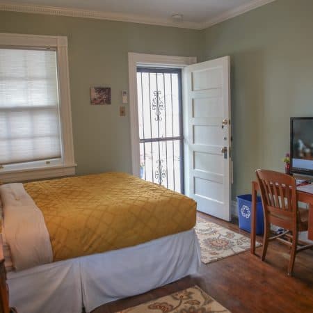 A bedroom with light green walls, a bed with a yellow comforter, a desk, and an open front door with a security screen.