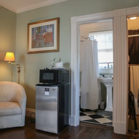 A bedroom with light green walls, a mini-fridge and microwave, and an open doorway leading to a bathroom with a black and white tiled floor.