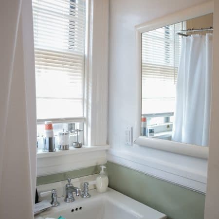 A bathroom corner with a sink, mirror, and window with blinds, showing a white shower curtain reflected in the mirror.