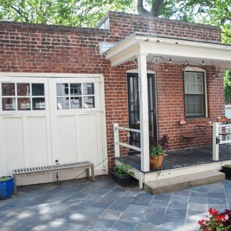 The exterior of a brick carriage house with white garage doors, a covered porch with string lights, and a patio with outdoor seating.