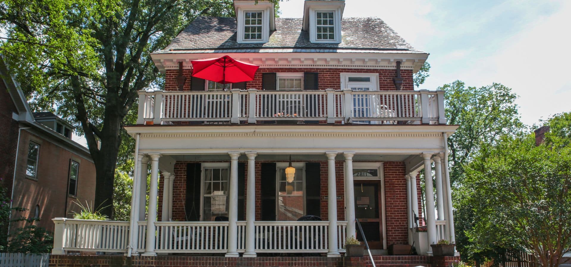 A two-story brick house with a red umbrella on the front porch, surrounded by trees.