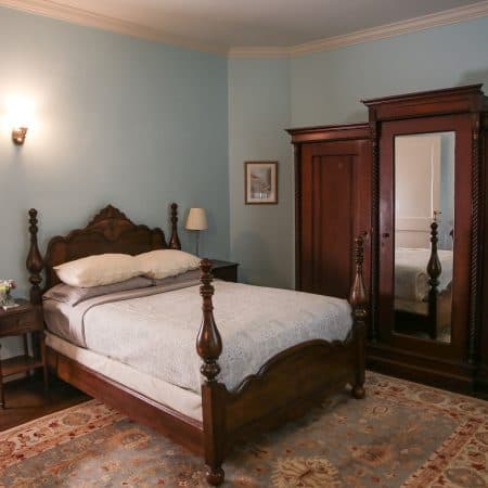 A bedroom with light blue walls, a dark wood four-poster bed, and a large wooden armoire.